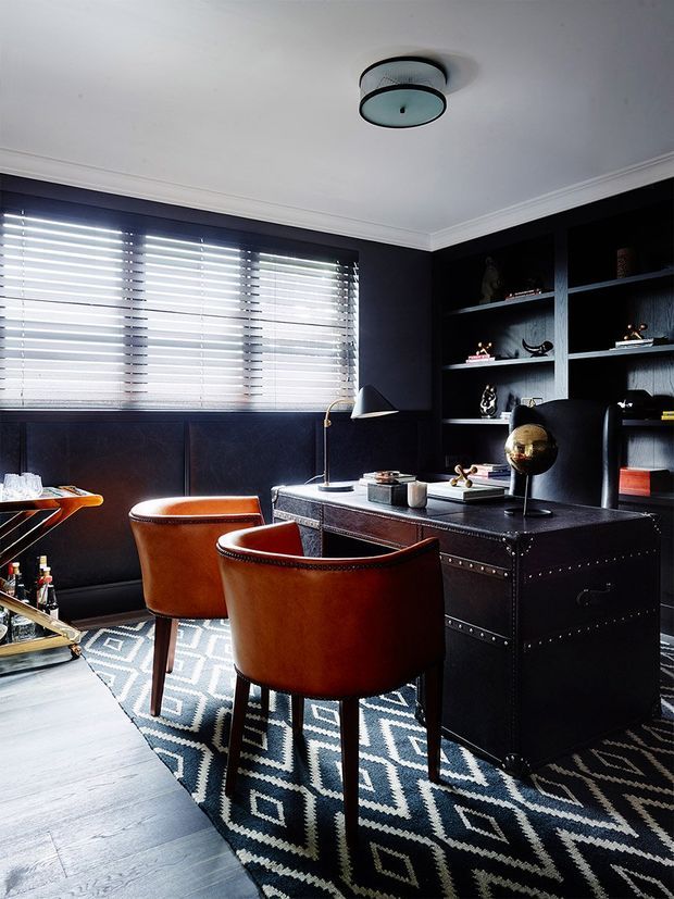 A sophisticated dark-toned home office with a black built-in shelving wall, paneled lower wall, and a substantial black desk. Two cognac leather chairs face the desk over a bold black-and-white geometric rug. Venetian blinds filter daylight through the wide window, while a sculptural desk lamp, decorative globe, and curated objects add refined character. White crown moulding and a contemporary ceiling light provide contrast.