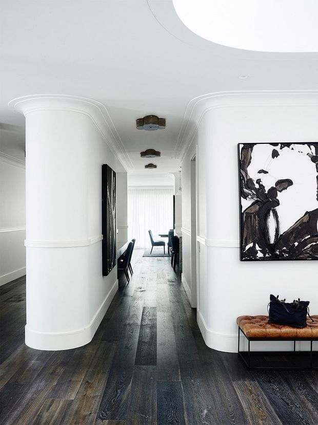 A bright, refined residential hallway with a dramatic dark timber floor, white curved architectural wall forms, decorative moulded cornices and wall trim, and a large circular ceiling opening or skylight. The corridor leads toward a glazed living or dining area, with contemporary artwork, dark seating, and a leather bench along the walls. The overall composition is monochromatic and elegant with strong contrast between the white walls and dark flooring.