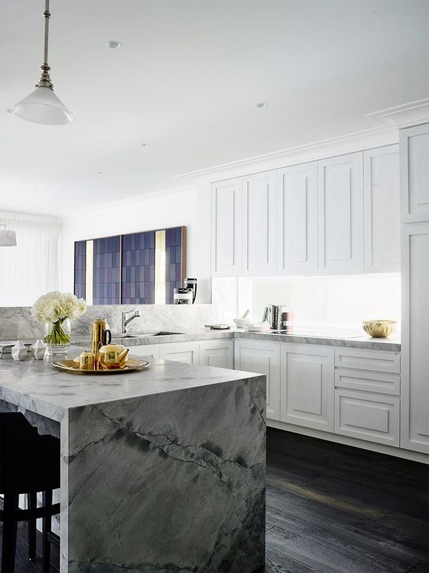 Bright, refined kitchen interior with extensive white Shaker-style cabinetry, a large dark veined stone island with seating, dark timber flooring, white pendant and recessed lighting, and a colourful geometric artwork. The space has a high-contrast white, grey, and dark-wood palette with subtle brass accents.