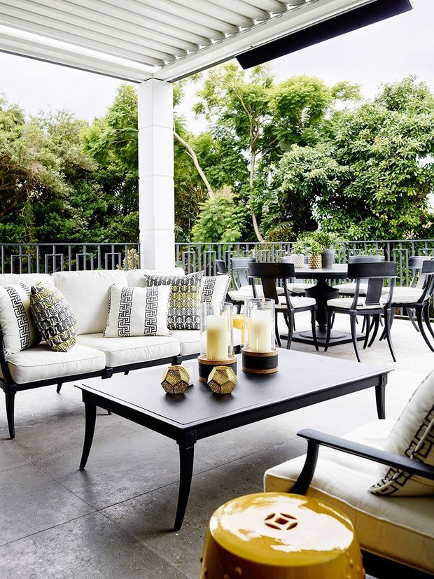 A bright covered outdoor terrace furnished as an alfresco lounge and dining area. The space has a large white upholstered sofa and armchair with patterned cushions, a black rectangular coffee table, matching black dining table and chairs, decorative candles, and a yellow side stool. A metal railing borders the terrace, with dense greenery beyond. The roof has exposed white ribbed metal panels and structural posts, while the floor is a smooth grey concrete or large-format slab surface.