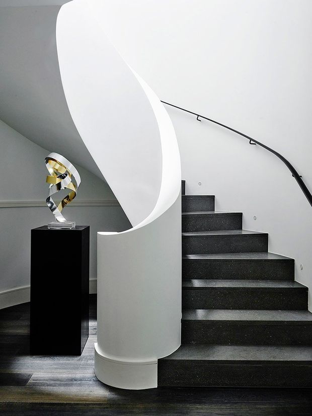 A sculptural interior stairway with dark stone or concrete steps, a sweeping curved white sculptural stair wall or balustrade, and a slim black handrail. A modern abstract metallic sculpture sits on a black pedestal beside the staircase, with dark timber flooring and light grey walls.