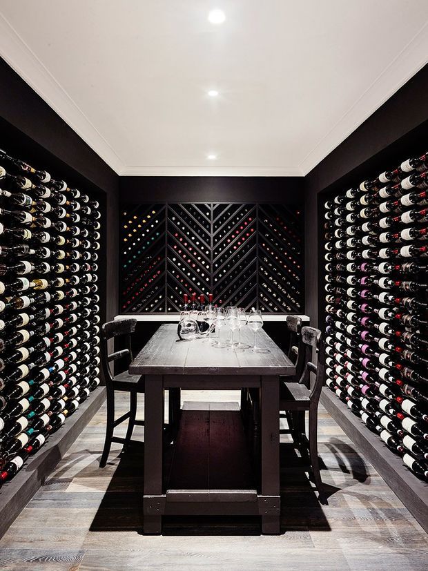 A compact, highly designed wine room with floor-to-ceiling racks of horizontally stored wine bottles lining both side walls and a decorative geometric bottle display at the rear. A dark timber or black dining-height table with chairs occupies the centre, set with wine glasses and decanters. The room has dark walls, a bright white recessed ceiling with downlights, and pale timber or stone-look flooring, creating a dramatic, luxurious atmosphere.