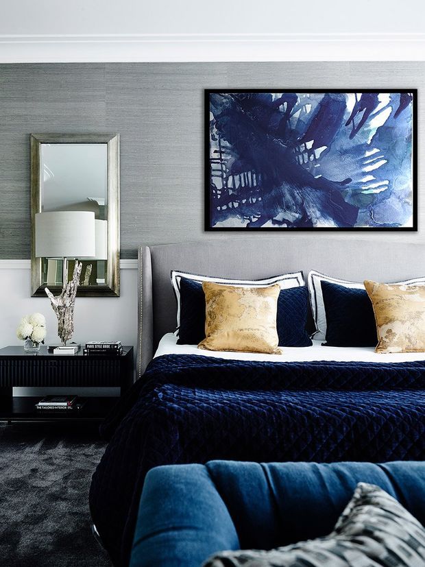 A refined bedroom interior featuring a large bed dressed in deep navy quilted bedding, navy and gold accent pillows, and a dark blue velvet seating piece in the foreground. The room has a grey textured feature wall with white decorative cornice, a rectangular metallic-framed mirror, black bedside furniture, a table lamp, and a bold blue abstract artwork. The overall look is dark, luxurious, and cohesive with navy, grey, black, and gold tones.