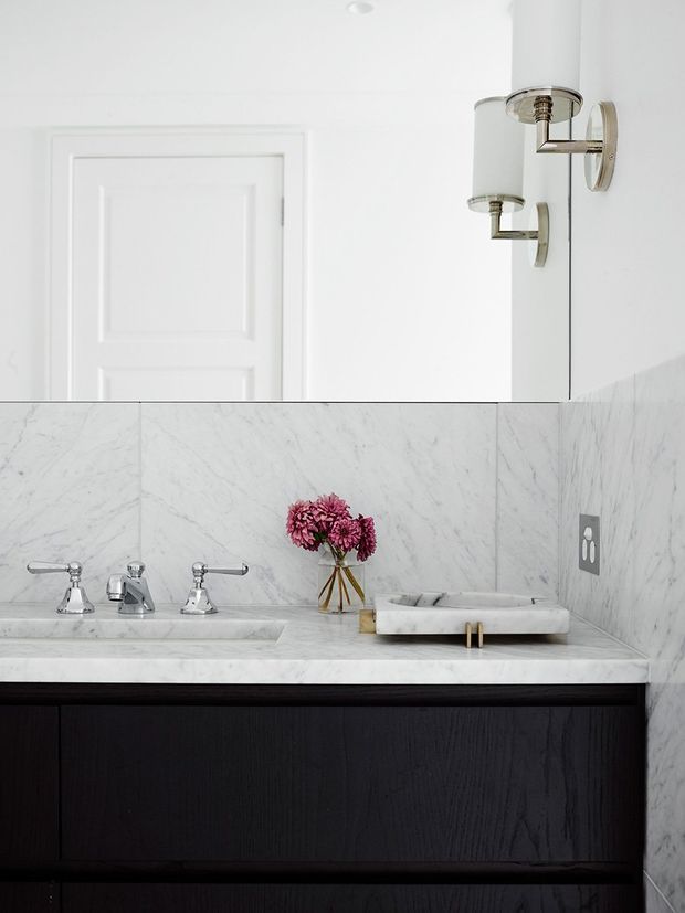 A refined bathroom vanity viewed straight on, featuring a white marble countertop and matching marble wall panels, a double-sink arrangement with polished chrome traditional taps, dark timber vanity cabinetry, and a white vessel basin with brass-toned supports. A large mirror, wall sconces, and a white paneled door complete the bright, high-contrast space.