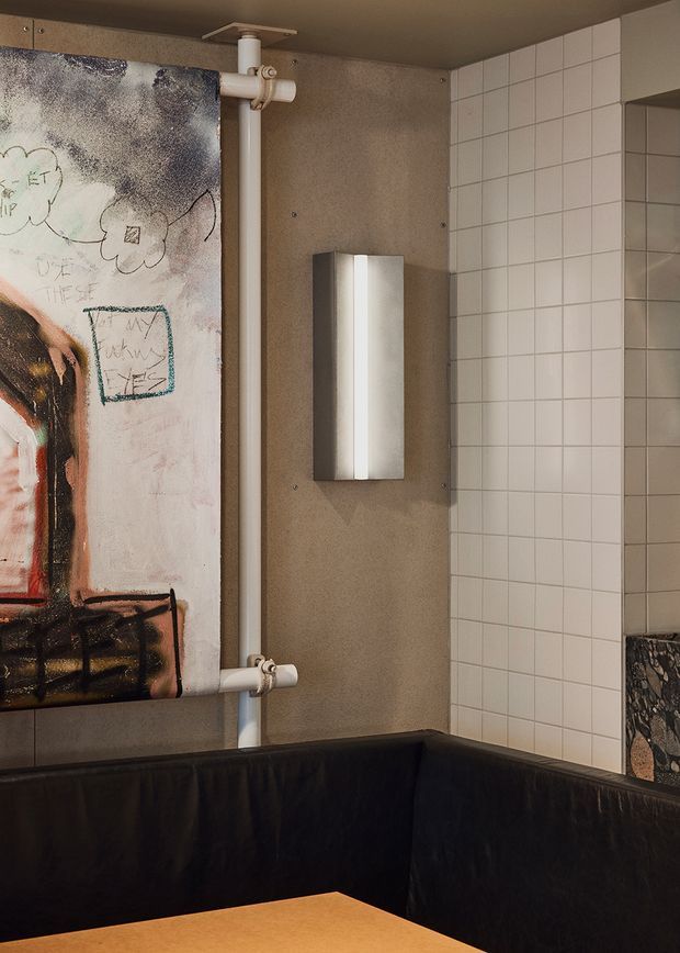 A moody, eclectic interior corner featuring a black upholstered booth or banquette, a warm wood tabletop, white square tiled wall surfaces, a rough taupe textured wall panel, exposed white pipework, a slim wall light, and a large expressive artwork mounted on a roller-style frame. A dark stone or marble surface is partially visible at the right.