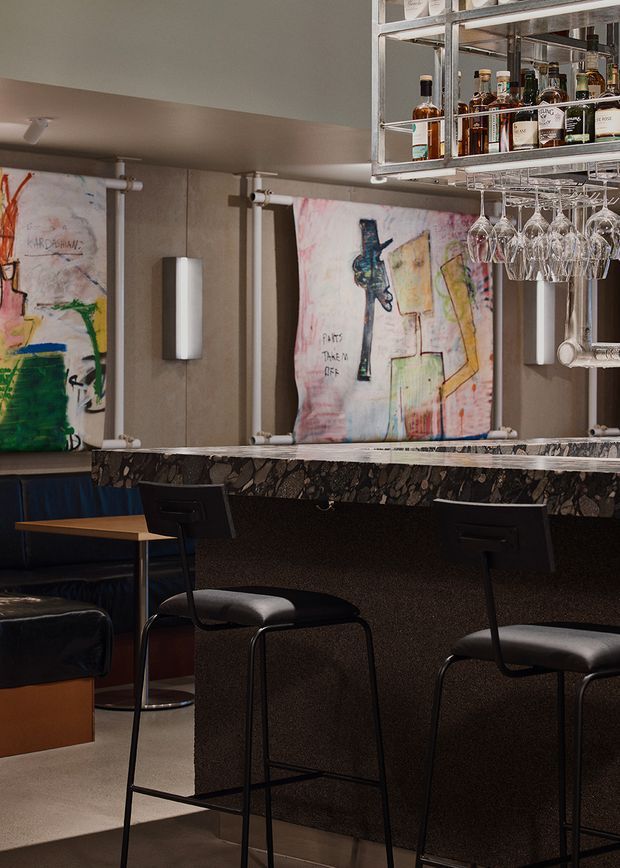 A moody contemporary bar interior with a dark stone-patterned counter, black bar stools, blue banquette seating, mirrored wall panels, exposed metal bottle shelving, and hanging wine glasses. Abstract artwork adds colourful visual accents to the otherwise dark, refined space.