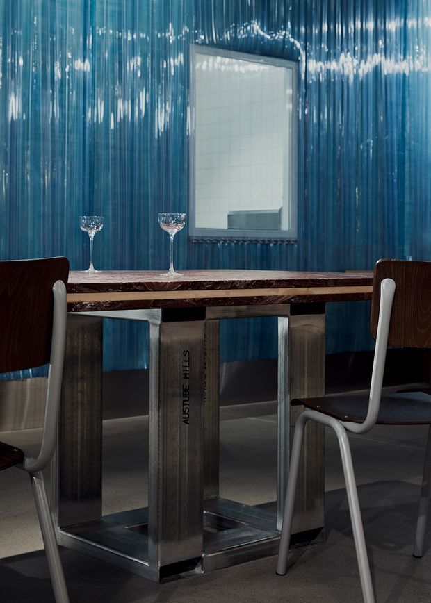 A compact, highly stylised dining area with a rectangular dining table featuring a dark marbled top and substantial brushed metal pedestal base. Two dark wood dining chairs with pale tubular frames flank the table. The walls are clad in glossy vertically ribbed blue material, with a rectangular mirror above the table reflecting a tiled surface. The floor is dark grey, and two wine glasses sit on the tabletop. The overall atmosphere is dramatic, moody, and luxurious.
