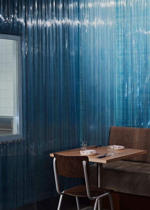 A moody restaurant or café dining booth with a small square timber table, upholstered banquette seating, and a single chair. The walls are covered in a highly reflective vertically ridged blue finish, creating a dramatic textured backdrop. A tiled service or kitchen area is partially visible through the opening at left.