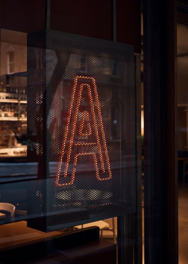A close-up view through dark-framed glazing of a perforated black metal sign or screen featuring a glowing orange letter A. Warm interior lighting and glimpses of upholstered seating, tables, and shelving are visible behind the screen, creating a dramatic hospitality or commercial interior atmosphere.