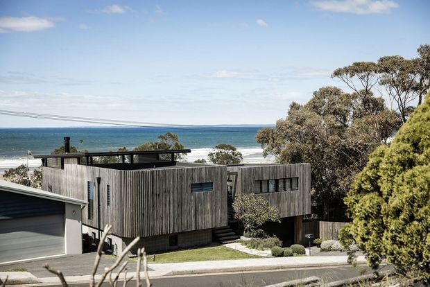 Exterior view of a contemporary coastal residence with dark weathered timber cladding, a flat roof, and strong horizontal and box-like forms. The home sits on a sloping suburban street overlooking the ocean, with mature coastal trees, a rooftop terrace or balcony, and an attached garage visible in the foreground.