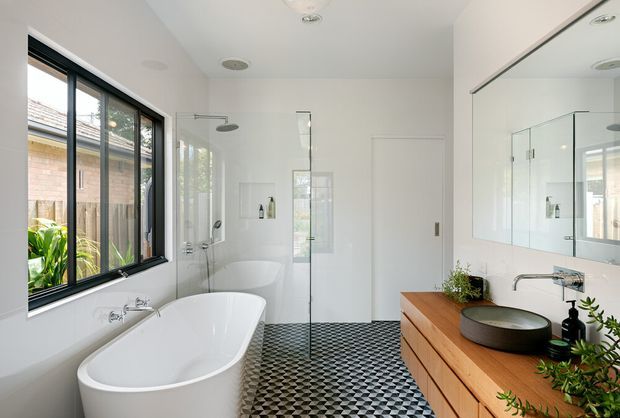 Bright contemporary bathroom with a freestanding white bathtub beside a large black-framed window, a frameless glass shower enclosure, wall niches, and a timber vanity with a round vessel basin. The room has white walls, a large mirrored cabinet, and a striking black-and-white geometric patterned tile floor.