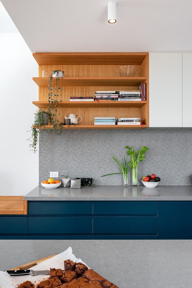 Contemporary kitchen view featuring deep navy-blue lower cabinetry, a grey stone benchtop, textured grey tiled splashback, and a warm timber open shelving unit with books and trailing plants. White upper cabinetry, a minimalist cylindrical ceiling spotlight, and a foreground island with brownies complete the clean, refined composition.
