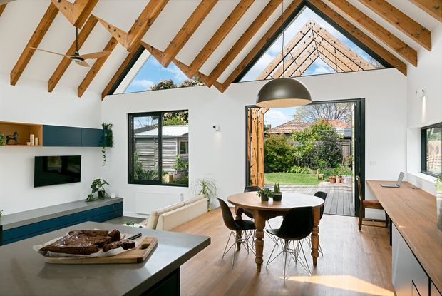 Bright open-plan living and dining area with a dramatic pitched ceiling, exposed timber beams and rafters, high triangular clerestory glazing, and large glazed doors opening to a garden patio. The room includes a modern kitchen island in the foreground, a timber dining table, built-in media cabinetry, timber flooring, and contemporary furniture.