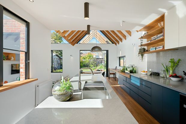 Bright contemporary kitchen with a large pale stone island and double-bowl sink, dark blue fixed cabinetry, timber flooring, open timber shelving, and a dramatic pitched ceiling with exposed timber rafters and a glazed triangular gable. Large windows provide abundant natural light and views into the garden.