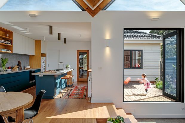 Bright open-plan residential interior showing a contemporary kitchen and dining area, with a dark blue island, light timber flooring, white cabinetry and walls, and a patterned runner. Large glazed doors open to a timber deck and courtyard, while a triangular timber-lined ceiling detail and skylight bring natural light into the space.