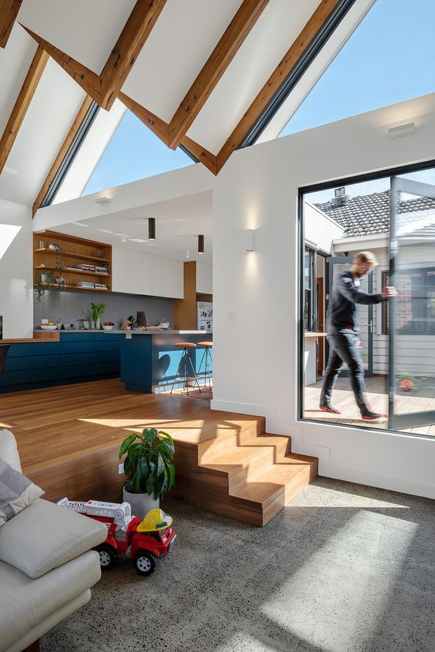 Bright open-plan residential interior combining a living area and kitchen, with a dramatic double-height ceiling, exposed timber roof beams, clerestory glazing, and large windows opening toward a courtyard. The space features polished speckled concrete flooring in the lower living zone, warm timber flooring and stepped timber platform, white walls, dark blue kitchen cabinetry, a grey stone-look splashback, timber shelving, and contemporary pendant and wall lighting. A child’s toy and sofa are visible in the foreground.
