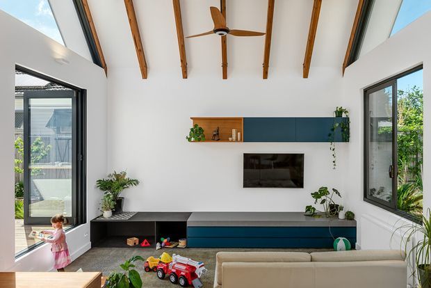 Bright contemporary living room with a high vaulted ceiling, exposed timber rafters, white walls, large black-framed windows and glazing, and a built-in dark teal and timber media cabinet. A wall-mounted television, indoor plants, sofa, ceiling fan, and children's toys are visible. The room opens toward an exterior courtyard or garden through large glazed doors.