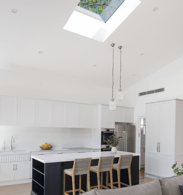 Bright, refined contemporary kitchen with extensive white shaker-style cabinetry, a dark island with open shelving, a pale stone benchtop, woven bar stools, stainless-steel appliances, pendant lights, and a large skylight. The room has a high ceiling and a light, neutral palette with dark cabinetry providing contrast.