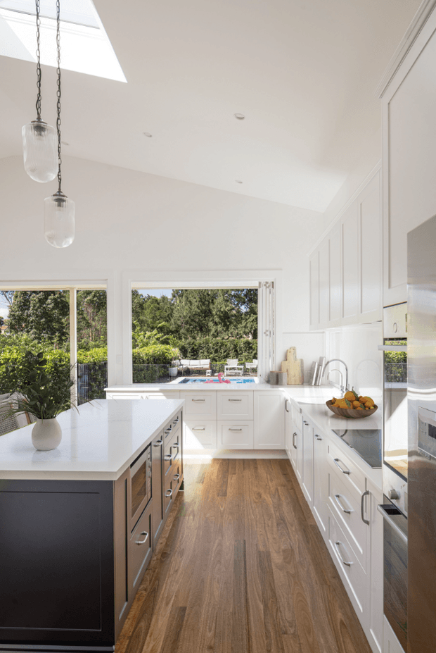 Bright, spacious white kitchen with a large central island, white shaker-style cabinetry, dark island joinery, stone benchtops, timber flooring, stainless-steel appliances, pendant lights, and generous windows overlooking a leafy outdoor area. A skylight adds abundant natural light.