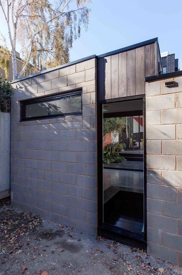 Exterior view of a compact contemporary addition or outbuilding with grey concrete block walls, dark metal-framed glazing and a vertically clad timber upper facade. A narrow glazed doorway opens toward an interior courtyard or garden, with a low, angular roofline and mature birch tree beside the structure.