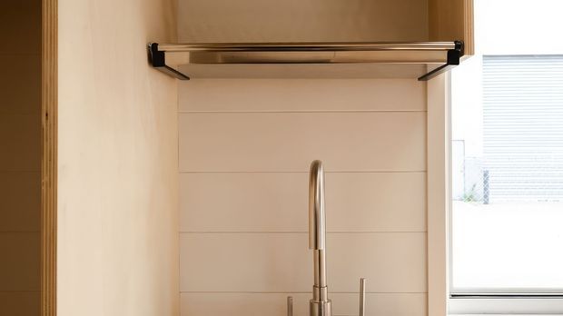 Close-up of a refined kitchen or wet-area joinery detail, featuring a tall brushed metal gooseneck tap, a horizontal rail or shelf above, pale timber-toned cabinetry, and a white horizontal-panel splashback beside a bright window.