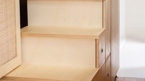 Close-up of built-in light timber cabinetry with open shelving and a fold-down or pull-out work surface. The joinery has visible layered plywood edges, pale wood grain, and dark hardware, with a woven panel partially visible at the left.