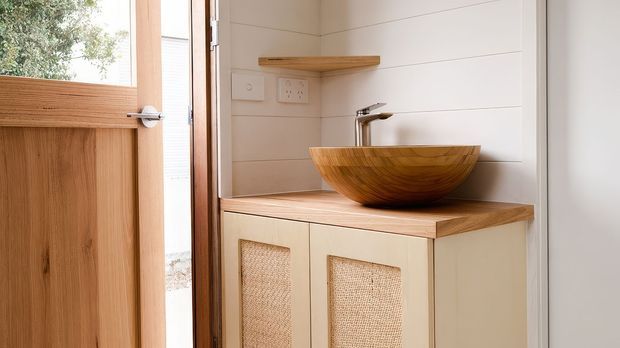 Compact washroom vanity area beside a timber-framed door, featuring a sculptural timber vessel basin on a timber benchtop, cream cabinetry with woven cane inserts, white horizontal wall panelling, and a small timber shelf. The space has a warm, natural, coastal-inspired fit-out with bright daylight.
