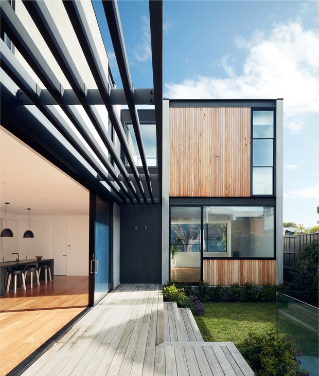 Modern two-storey residential courtyard facade viewed from a timber deck. The home combines dark metal framing, extensive glazing, and vertical timber cladding, with a dramatic pergola of deep horizontal slats extending over the deck. An open-plan kitchen and dining area is visible through the large sliding glass doors, while the courtyard includes lawn, planting, fencing, and a glass balustrade.