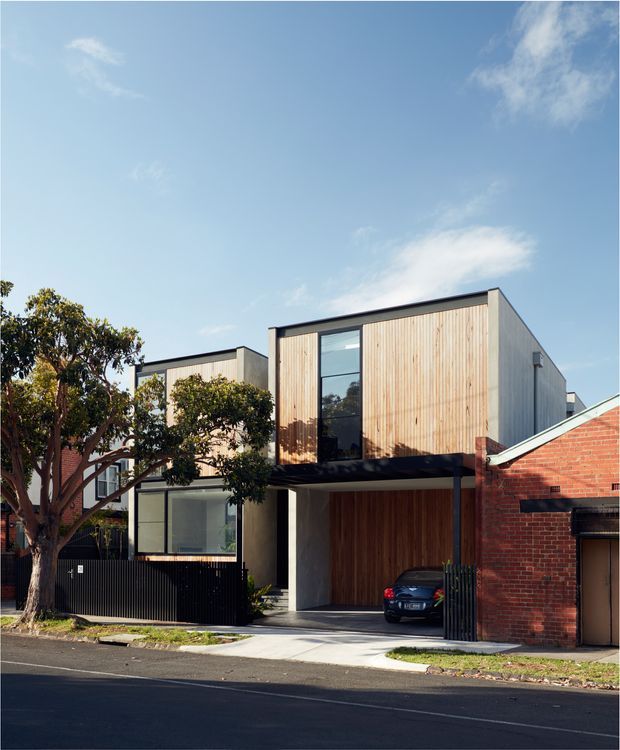 Street-facing view of a contemporary multi-storey urban residence, combining light vertical timber cladding, dark metal-framed glazing and a projecting upper level over a sheltered carport. A red-brick neighbouring building is visible at the right, with established street trees and a paved driveway in front. The design has clean rectilinear massing and a flat roofline.