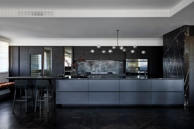 A sophisticated dark-toned kitchen with a large central island, integrated cabinetry, black marble splashback and side wall, dark herringbone flooring, and a sculptural multi-light pendant. Bar stools sit at the island, while reflective cabinetry and glazing add depth. A decorative linear ceiling cornice provides a refined architectural detail.