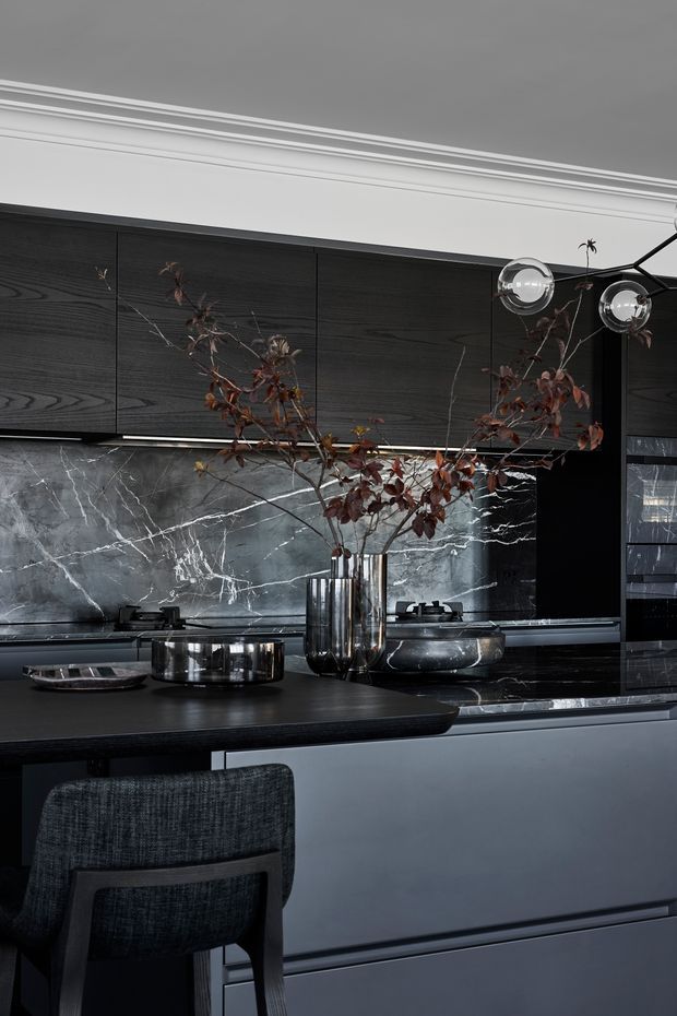 A sophisticated dark-toned kitchen with black and charcoal cabinetry, a dramatic grey marble splashback, dark benchtops, and a large island with integrated seating. Metallic pendant lights, polished metal cookware, and a tall vase of dried branches add reflective and organic accents. The space has a refined, moody, high-contrast aesthetic with classic ceiling moulding.