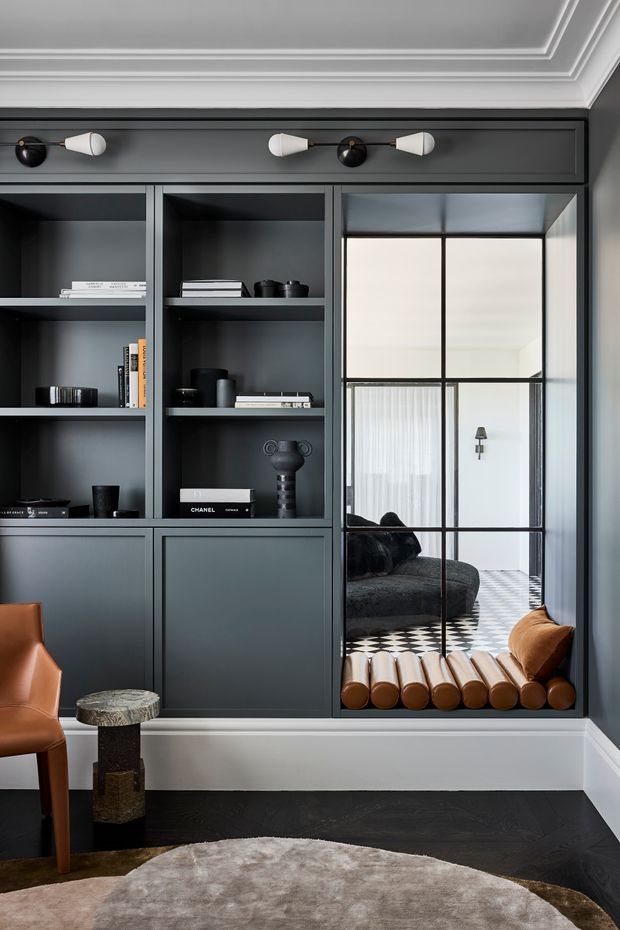 A refined, dark-toned interior featuring built-in charcoal cabinetry and open shelving, a mirrored steel-framed panel, decorative wall lighting, and classic ceiling mouldings. The reflection reveals a lounge area with a dark sofa, sheer curtains, and a black-and-white checkerboard floor. A warm leather chair, sculptural side table, and round neutral rug add contrast to the moody joinery.