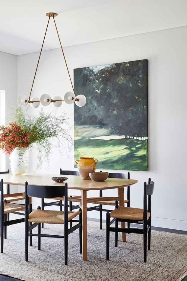 A bright, refined dining room with a rectangular timber dining table surrounded by black-framed woven-seat chairs. A sculptural linear pendant with white globe shades hangs overhead, while a large dark landscape artwork provides a dramatic focal point against pale walls. A textured neutral rug, ceramic vessels, and a floral arrangement complete the space.
