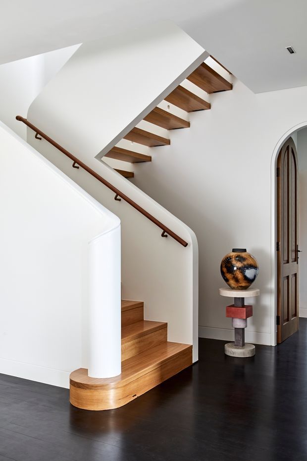 A refined interior stairway with warm timber treads and exposed timber stair soffit, set against smooth white curved walls and a sculptural white balustrade. A dark timber handrail follows the stair, while a tall arched timber doorway and decorative pedestal feature in the adjacent hall. The room has dark flooring, clean detailing, and abundant natural light.