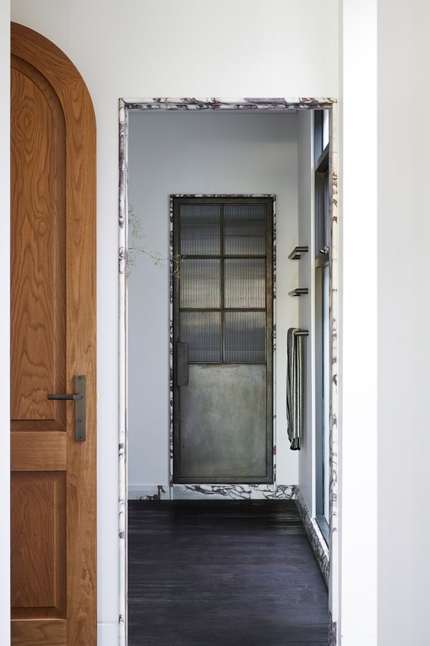 A narrow, contemporary entryway or hallway with a dark timber floor, white walls, and a dramatic marble-patterned door surround and skirting. A tall industrial-style metal door with ribbed glass panels and a weathered metal lower panel anchors the far end. A large curved-top timber door is partially visible on the left, while a glazed opening and dark metal wall hooks or handles appear on the right.