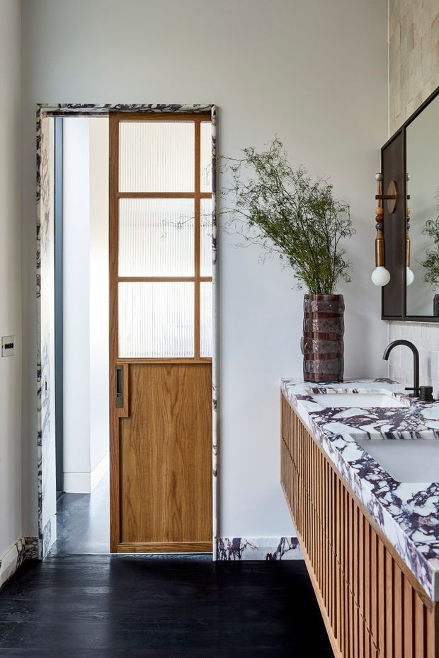 A refined bathroom interior featuring a floating timber vanity with vertical slatted cabinetry, a dramatic veined stone benchtop and integrated basin, black tapware, and a large dark-framed mirror. A timber door with reeded glass opens to a tiled adjoining space. The room combines warm wood, pale walls, dark flooring, and sculptural decorative elements.