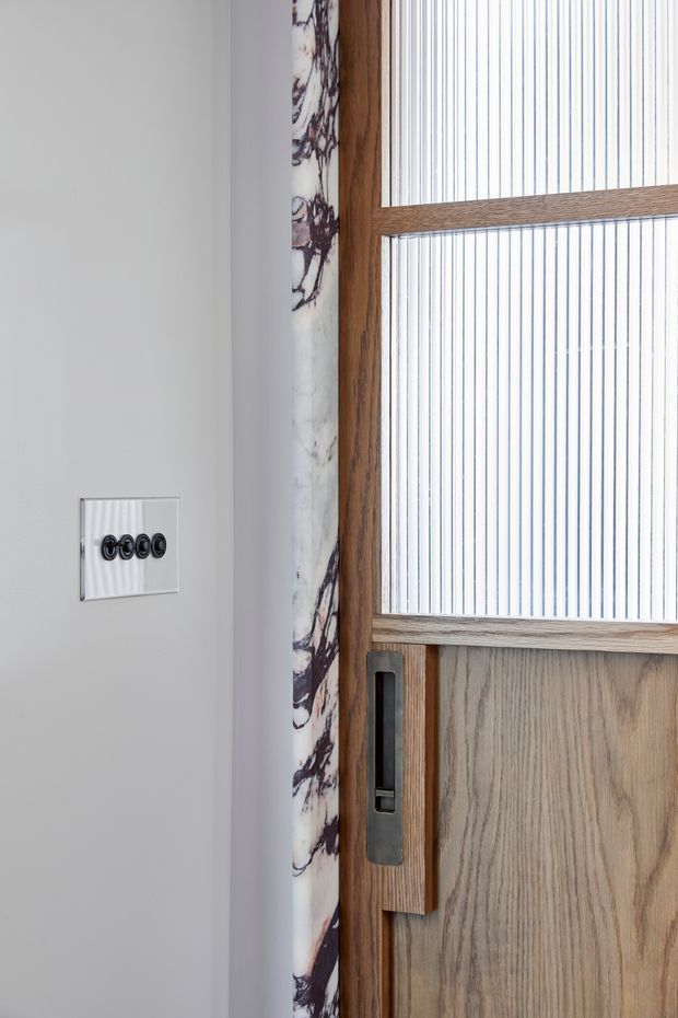 Close-up interior detail of a timber-framed door with vertically reeded glass panels and a dark metal recessed pull handle. A white wall with a modern multi-button light switch is visible beside the door, along with a narrow veined marble trim.
