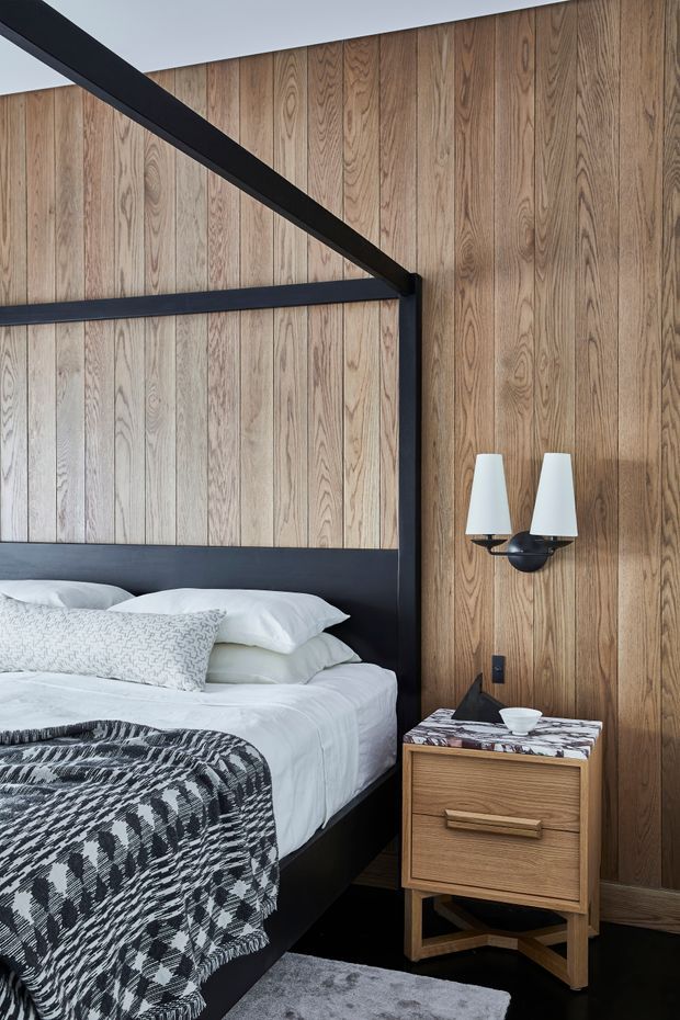 A refined bedroom with a black-framed four-poster bed, white bedding, and a black-and-white patterned throw. The room features full-height vertical timber wall panelling, dark flooring, a sculptural black wall sconce with twin white shades, and a compact timber bedside table with a stone or patterned top. The overall design is warm, contemporary, and high-contrast.