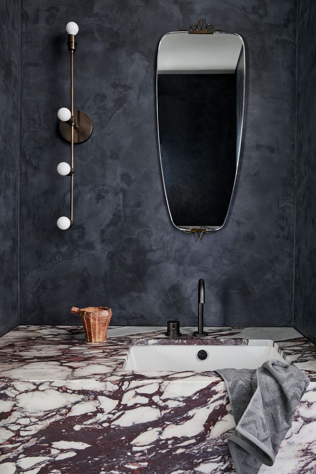 A dramatic, moody bathroom vanity featuring a white inset basin set into a richly veined burgundy-and-white marble benchtop. The walls are finished in dark charcoal textured plaster, with a slim black tap, round black controls, a vintage-inspired vertically oriented mirror, and a sculptural wall light with globe bulbs. A grey hand towel and copper-toned vessel add contrast.
