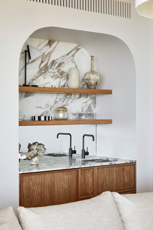 A refined built-in bar and kitchenette niche viewed behind a sofa. The arched alcove has dramatic white marble veining on the backsplash and benchtop, warm timber open shelving and cabinetry, matte black twin taps, an integrated sink, and carefully styled glassware and ceramic vessels. A linear ventilation grille is visible above, with soft neutral walls and upholstery creating a warm, contemporary setting.