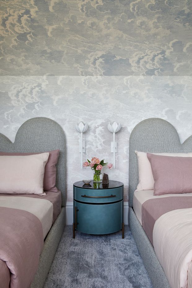 A softly styled bedroom with two single beds positioned symmetrically on either side of a round blue bedside table. The beds have tall rounded upholstered grey headboards and layered blush, mauve, and cream bedding. The walls and ceiling are covered in a pale blue-grey and cream floral cloud-patterned wallpaper, with white wall-mounted decorative lights between the beds. A blue-grey rug and small floral arrangement complete the calm, refined scheme.