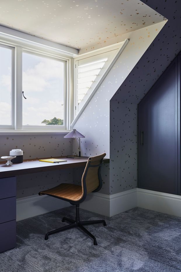 A compact, tailored home office in a dormer-like alcove, with a built-in dark desk beneath a large corner window, white plantation shutters, a mid-century style wood-and-black office chair, and a small pleated desk lamp. The walls, sloped ceiling, and deep blue built-in cabinetry are covered in a whimsical star-patterned wallpaper, while grey carpet and white skirting create a soft, calm base. Natural daylight fills the room.