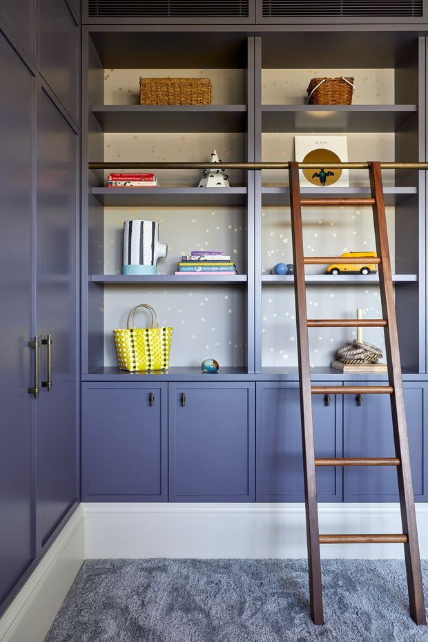 A built-in children's room storage wall in deep blue cabinetry, with open shelving, decorative wallpaper, baskets, books and toys. A timber library ladder leans against the shelving, and grey carpet covers the floor. The space has a playful yet refined traditional built-in joinery design.
