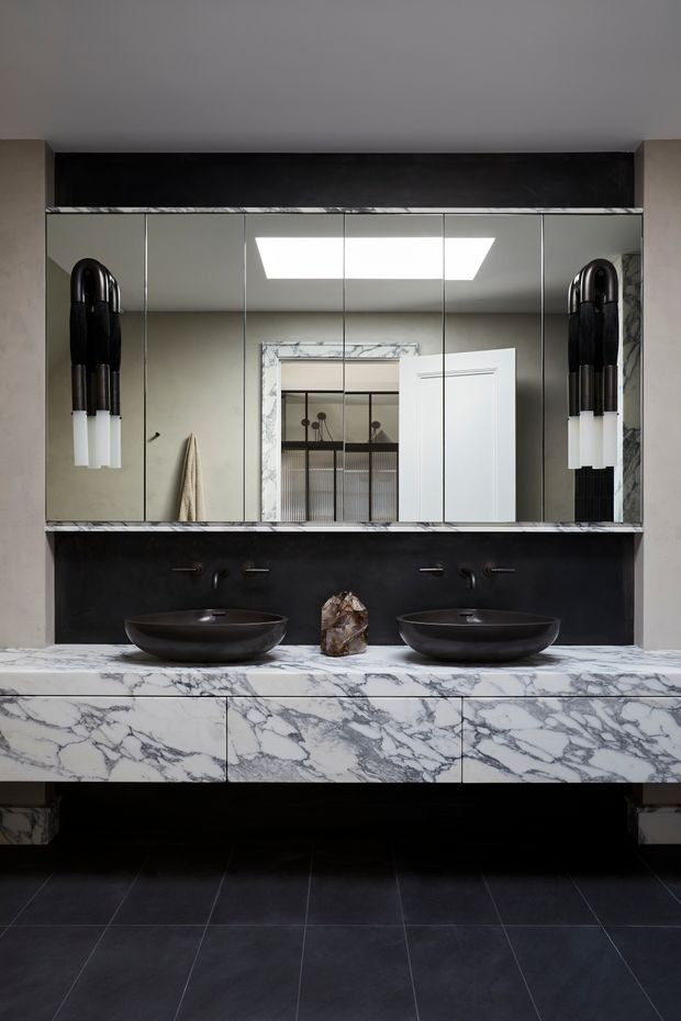 A sophisticated double-vanity bathroom with two dark vessel basins set on a dramatic white-and-grey veined marble vanity. The wall behind the basins is clad in matte black, with a large mirrored cabinet above and sculptural black wall sconces with white cylindrical shades on either side. Dark floor tiles, pale neutral walls, and a reflected skylight create a refined, high-contrast interior.