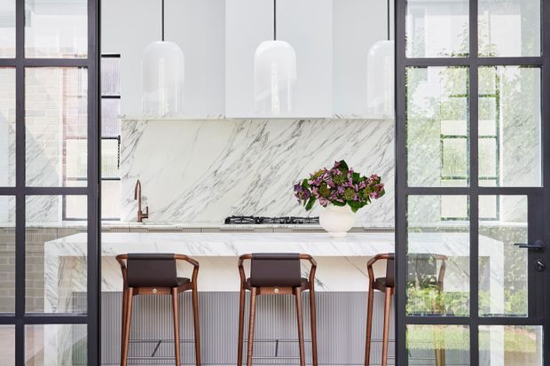 Bright contemporary kitchen viewed through black-framed glazed doors. The space features a large white marble island with waterfall edges, matching marble backsplash, white cabinetry, ribbed island detailing, three bar stools, dark-framed glazing, and sculptural white pendant lights. A floral arrangement adds colour to the otherwise refined neutral scheme.