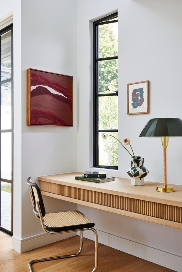 A bright, refined home office nook with a wall-mounted timber desk featuring vertically fluted detailing, a cantilevered cane-and-black chair, warm timber flooring, tall black-framed windows, and curated artwork and decorative objects. The neutral white walls and natural light create a clean, contemporary workspace.