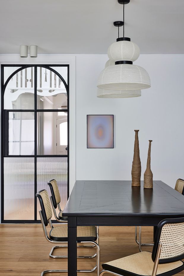 A refined dining room with a large dark rectangular dining table surrounded by black-framed cane and chrome cantilever chairs. Sculptural woven pendant lights hang above the table, while a black-framed glazed partition with arched detailing opens toward a bright adjoining space. Light timber flooring, pale walls, and understated artwork create a calm contemporary setting.