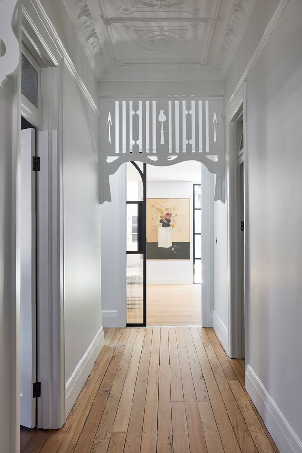 A narrow, bright residential hallway with restored ornate ceiling plasterwork, decorative wall and doorway mouldings, white-painted walls and trim, and aged timber floorboards. An elaborate white fretwork screen frames the opening to a light-filled room beyond, with a black-framed glazed door and artwork visible in the adjoining space.