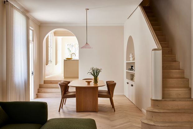 Warm, refined open-plan dining and living area in a renovated-looking home, featuring a round timber dining table with leather chairs, pale herringbone timber flooring, soft white walls, arched openings and built-in display niches. A curved timber staircase rises beside the dining area, while tall sheer window coverings bring in diffused daylight. A partial green sofa is visible in the foreground and a compact kitchen or utility area appears beyond the archway.