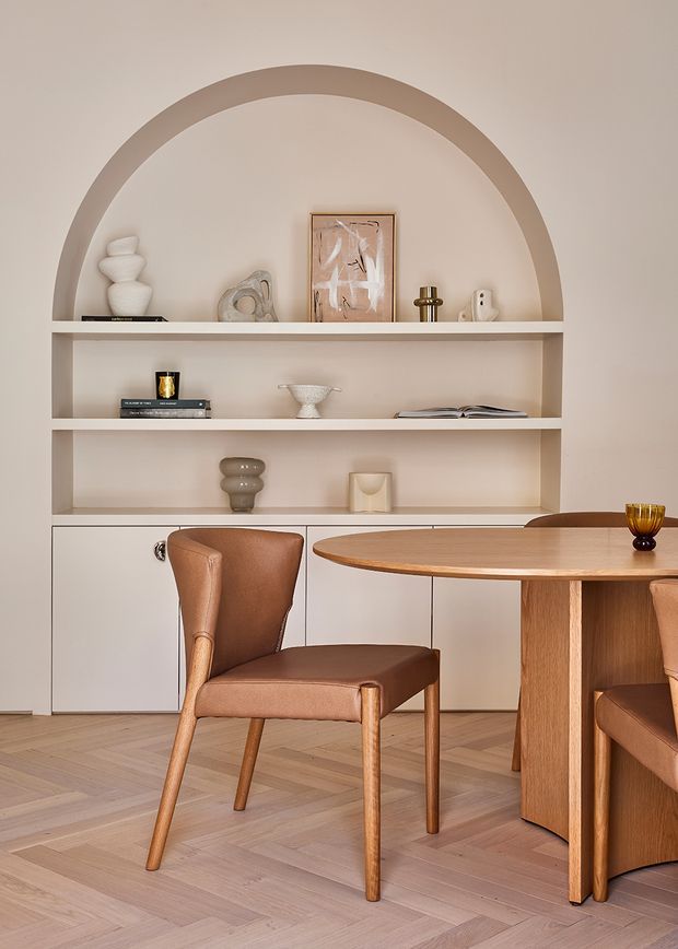 Warm, refined dining area featuring a round timber dining table and leather-upholstered chairs in front of custom built-in shelving. The shelving incorporates a large arched alcove and open display niches with neutral decorative objects, while pale herringbone timber flooring and soft beige finishes create a cohesive, minimalist interior.